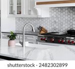 A kitchen sink detail, stainless faucet on a white farmhouse sink and marble counter, and a marble hexagon tiled backsplash with white cabinets. No brands or labels.