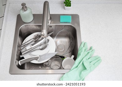 A kitchen sink piled with dirty dishes awaiting cleaning, accompanied by dishwashing soap, a sponge, and a pair of green rubber gloves on the countertop. - Powered by Shutterstock