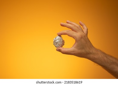 Kitchen Tin Foil Crumpled Into A Ball Held In Hand By Caucasian Male Hand. Close Up Studio Shot, Isolated On Orange Background.