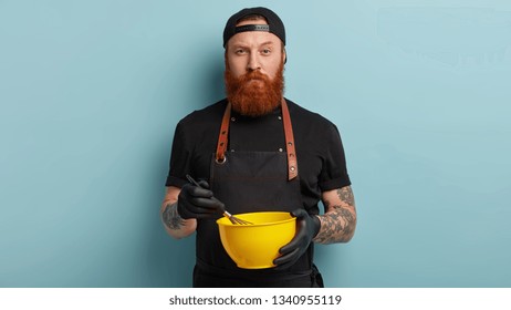 Kitchen Tools And Cooking Concept. Serious Bearded Man Restaurant Worker Uses Whisk And Bowl For Making Cream, Mixes Milk With Eggs And Sugar, Has Tattoo On Arms, Stands Over Blue Background.