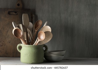 Kitchen utensil set in rough handmade ceramic pot, crockery, cutting boards on wood table against rustic wooden wall. Low light.
