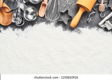 Kitchen utensils and flour for baking or cooking background. Rolling pin, whisk, wooden spoon, cookie cutters and measuring spoons on dark grey concrete background with copy space for text. Top view.