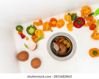 Kitchen vegetable scraps and eggshells are scattered around a white sink under bright natural light. The image shows sliced peppers, eggshells, and food remnants near the drain. The setting captures a