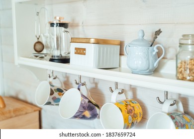 Kitchen wooden shelf with tea leaves in gold box and accessories, blue sugar bowl with spoon, strainer, press. Many colorful cups, mugs are hanging from hooks. Cozy interior in a country house.