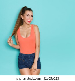 Kittenish Girl Pulling Her Hair. Sexy young woman in orange shirt with cleavage pulling ponytail and smiling. Three quarter length studio shot on teal background.