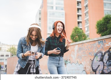 Knee figure of two young handsome caucasian blonde and redhead straight hair women dancing in the city listening music with headphones and smartphone, laughing - music, fun, freedom concept - Powered by Shutterstock