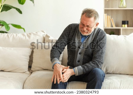 Knee pain arthritis body sick health care concept. Unhappy middle aged senior man suffering from knee ache sitting on sofa at home. Mature old senior grandfather touching leg feeling pain hurt in knee