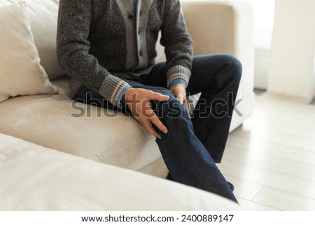 Knee pain arthritis body sick health care concept. Middle aged senior man suffering from knee ache sitting on sofa at home. Mature old senior grandfather hands touching leg feeling pain hurt in knee