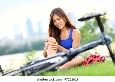 Knee Pain Bike Injury. Woman With Pain In Knee Joints After Biking On Bicycle. Girl Sitting Down With Painful Face Expression. Mixed Race Sport Fitness Model Outdoors.