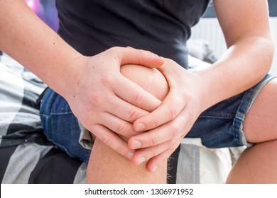 Knee pain concept photo. Caucasian male holding both hands behind knee, which pierced acute sharp pain while sitting on edge of sofa. Symptom of diseases of joints, ligaments, and cartilage of knee