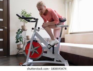 Knee problems. Young adult athletic woman holding on to her knee and writhing in pain, she was injured while riding a home exercise bike against the backdrop of a sofa and home plants - Powered by Shutterstock