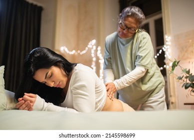 Kneeling pregnant woman and midwife at home. Woman in casual clothes leaning on bed, Asian doula massaging back. Pregnancy, medicine, home birth concept