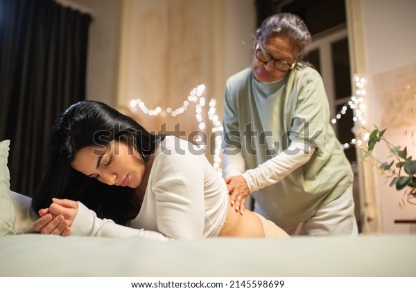 Kneeling pregnant woman and midwife at home.
Woman in casual clothes leaning on bed, Asian doula massaging back.
Pregnancy, medicine, home birth
concept