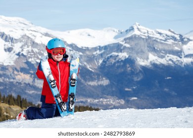 Kneeling in the snow, a happy youngster in a red jacket grips a pair of skis with majestic snow-covered peaks in the background - Powered by Shutterstock