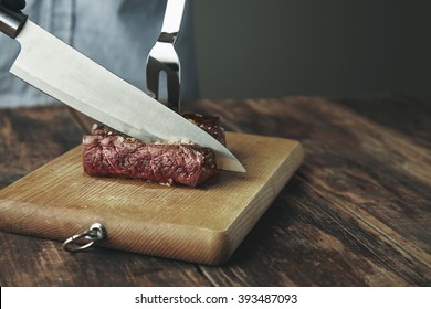 Knife Cut Slice Of Grilled Meat On Wooden Board In Front Of Big Steel Fork In Steak. All Isolated On Side Of Vintage Table