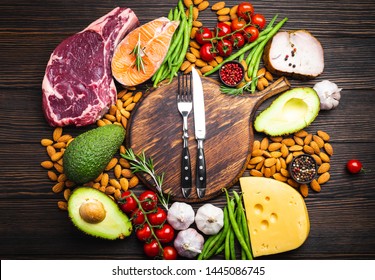Knife and fork over wooden cutting board and ketogenic low carbs ingredients for healthy eating concept and weight loss, top view. Keto foods: meat, fish, avocado, cheese, vegetables, nuts