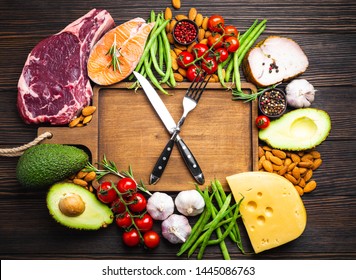 Knife And Fork Over Wooden Cutting Board And Ketogenic Low Carbs Ingredients For Healthy Eating Concept And Weight Loss, Top View. Keto Foods: Meat, Fish, Avocado, Cheese, Vegetables, Nuts