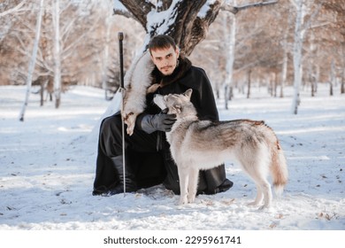 a knight in a black cloak and in furs with a sword in his hand in a snowy forest. Young male warrior in winter with a wolf. A guy with a Siberian helmet. fantasy image