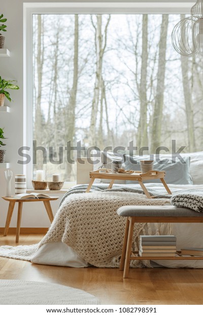 Knit blanket on bed next to wooden table against the fake window bedroom wallpaper interior decoration. 