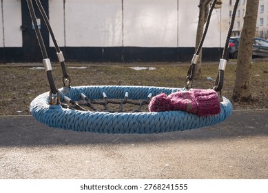 Knitted hat left on an empty rope swing in a playground during mild spring weather, illustrating a seasonal transition and the common occurrence of misplaced personal items