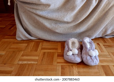 Knitted Home Slippers Shoes On Wooden Floor Near Sofa Covered With Blanket. Conceptual For Real Life Moments