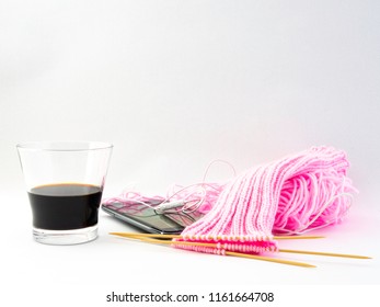 Knitting a pink pair of socks while drinking coffee and having the phone nearby. Tabletop front view with a white background. Pink wool on the table and wooden double pointed knitting needles. 