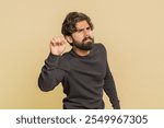 Knock-knock who is there. Confused Indian man knocking door gesture asking who is at home, feeling embarrassed, no idea, being clueless and uncertain. Young guy isolated on beige studio background.