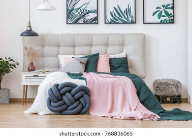 Knot pillow and green and pink bedding on bed in cozy bedroom interior with lamps, posters and suitcase