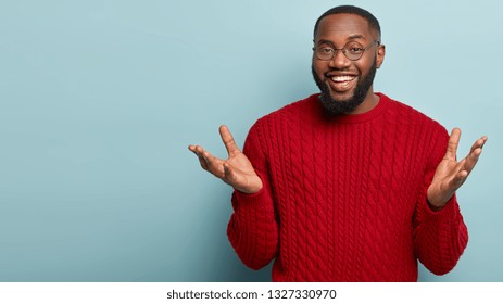 I know nothing. Horizontal shot of friendly looking black man spreads hands, gestures actively while explains something to interlocutor, stands over blue background, mock up space aside, has no idea - Shutterstock ID 1327330970