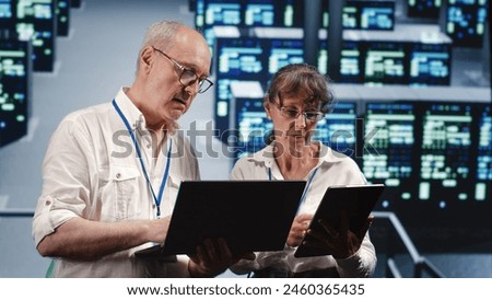 Knowledgeable IT practitioners looking around server farm, using laptop and tablet to crosscheck disaster recovery plan and assess rackmounts in need of replacement, preventing perils