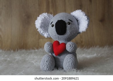 Koala Bear Hold A Little Red Heart. Grey Knitted Koala Bear Toy Sitting On Fluffy White Blanket, Wooden Background. Valentine Concept Photo Idea