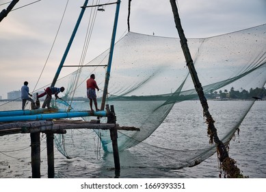 Kochi, India - December 24, 2019: Men Trying To Take Out The Fish From Chinese Fishing Nets Above Water, Near The Empty Shore In The Afternoon, In Kochi, India.