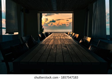 Koh Samui / Thailand - November 16, 2020 :  Room on the beach with a long wooden table with chairs against the backdrop of an open window at a spectacular colorful sunrise over the sea