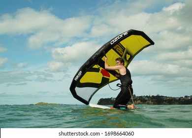 Kohimarama, Auckland / New Zealand - October 18 2019: A young man wingfoils in Auckland harbour, using a hand held inflatable wing and riding a hydrofoil surfboard.  Green sea and cloudy blue sky.