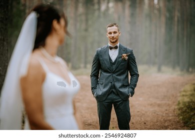 Koknese, Latvia - September 26, 2023 - Groom in a tailored suit stands confidently in a forest, gazing at the bride in soft focus, creating a romantic outdoor wedding atmosphere. - Powered by Shutterstock