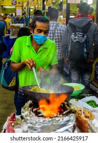 Kolkata, India - February 13, 2022: A Man Tossing Noodles In A Black Pan With Sizzling Smoke Coming Up. People Gathered In The Background To Enjoy The Food Festival.