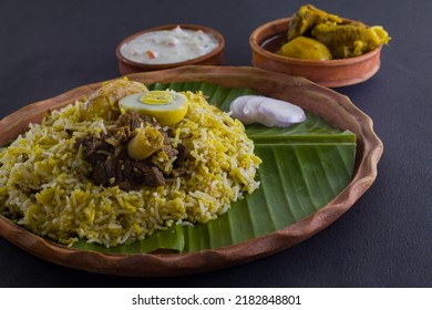 Kolkata Style Mutton Biryani With Potato And Egg Served On Clay Plate And Banana Leaf With Mutton Curry And Curd Raita. Shot Against Black Background. 