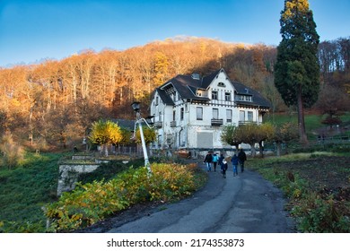 Konigswinter, Germany - November 28, 2020: People Walking On A Path In Front Of An Old White House With A Light Post Falling Over In Front Of A Colorful Hill With Light Shining On The Trees.