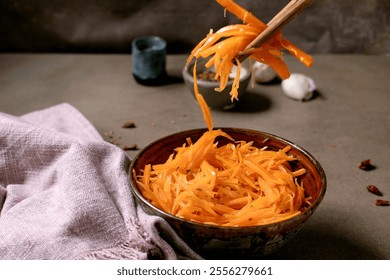 Korean and Asian cuisine. Man's hand uses chopsticks to pick up vegetable salad of marinated carrots with garlic from ceramic bowl. Seasoning in small bowls on Mocha Mousse tones table. Healthy eating - Powered by Shutterstock