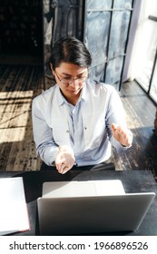 Korean Businessman In A Shirt Sits At A Table And Works On A Laptop, Gesturing And Communicating With Workers Via A Video Call