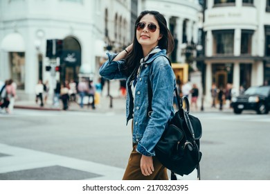 Korean Lady Standing On Street Is Combing Hair With Hand And Looking Back With A Charming Smile. Fashionable Woman Carrying A Backpack Is Walking In The City With Romanesque Architecture. 