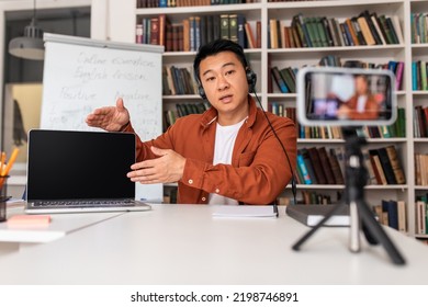 Korean Male Teacher Teaching Online Having Remote Lecture Showing Laptop Blank Screen Filming Lesson On Cellphone Sitting At Workplace Indoor. Selective Focus, Mockup