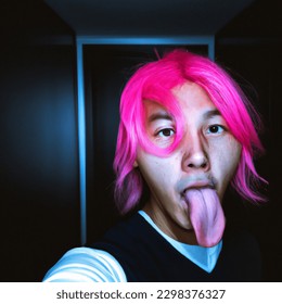 Korean Man with blue eyes and pink hair sticking his tongue out inside a Dark room