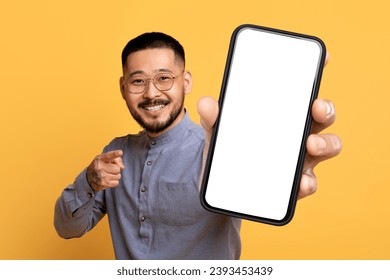 Korean man with large blank cellphone pointing to camera, showing mobile touchscreen perfect for internet and chat app mockups, studio shot over bright yellow backdrop, modern gadget concept - Powered by Shutterstock