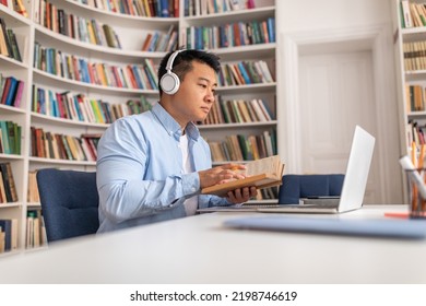 Korean Middle Aged Male Learning Online Holding Book And Using Laptop Computer Wearing Headphones Sitting In Modern Library. Side View Of Adult Learner Having Online Lecture