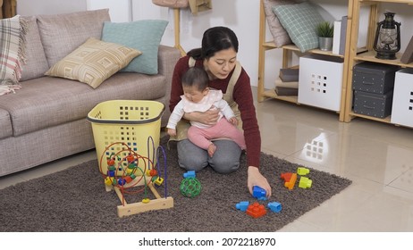 Korean Mom Kneeling On The Carpet Is Picking Up Toys Into The Basket With Her Toddler Child In Arms At Home Living Room.