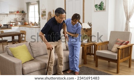 korean woman in-home care attendant assisting senior male to stand up from the sofa at home. the senior patient using a crutch is feeling great pain in his knee joints