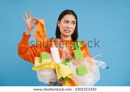Korean woman looks at stinky latex glove, holds plastic garbage, cleans house and recycle, blue background.