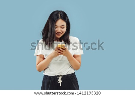 Korean young woman's half-length portrait on blue studio background. Female model in white shirt. Chating on the phone. Concept of human emotions, facial expression. Front view.