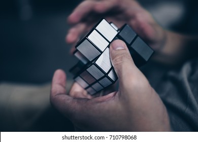 Koronadal City, PH – September 1, 2017: Rubik's Cube Was Invented In 1974 By Erno Rubik. A Man’s Hand Trying To Solve The Irregular Silver Puzzle With Dark Background. Side View. Selective Focus.